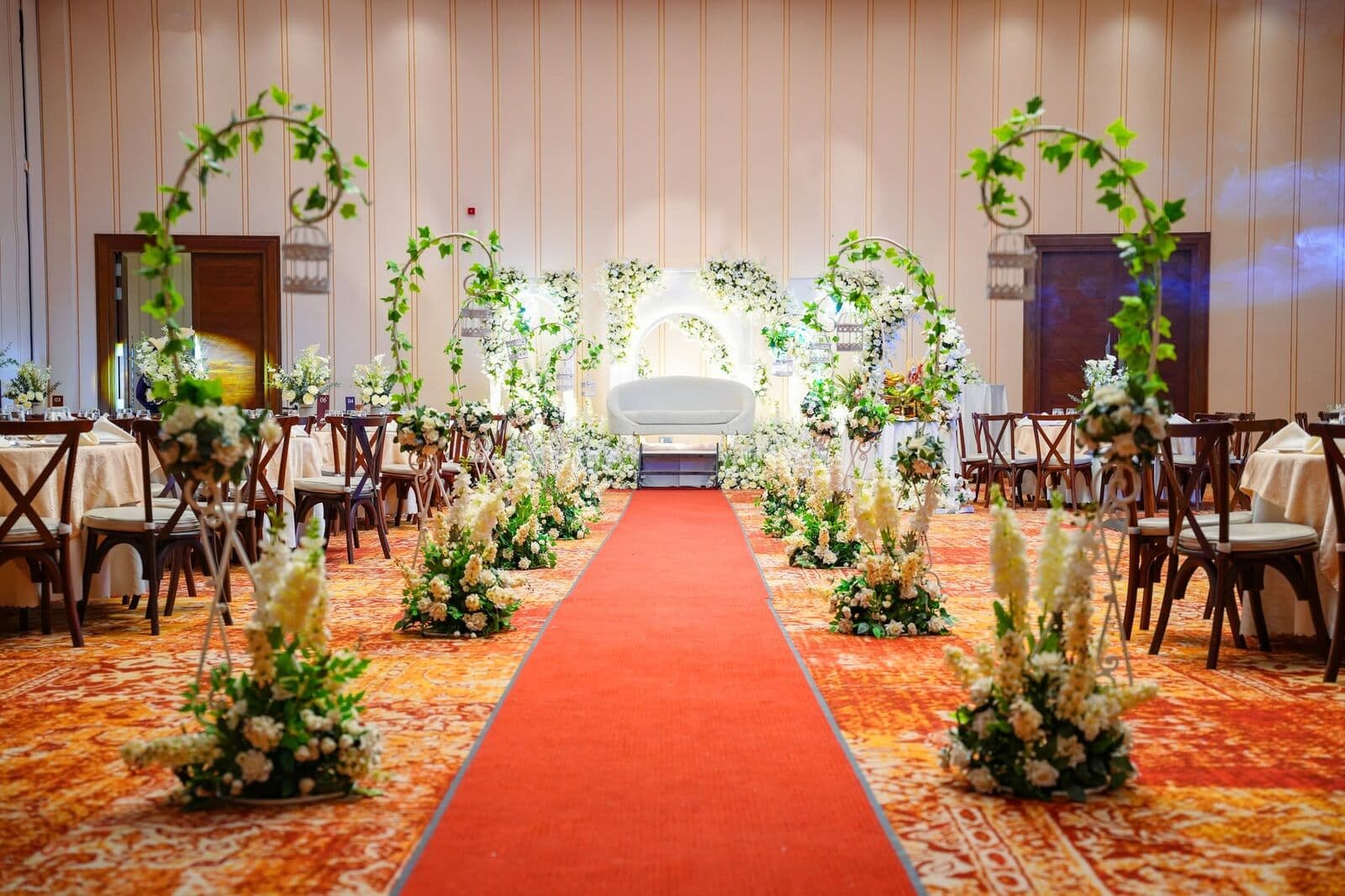 A red carpet aisle lined with white flowers and green arches, leading to the couple's seat