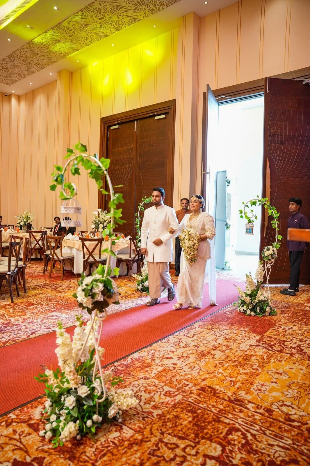A bride and groom walking into the hall along a red carpet lined with flowers