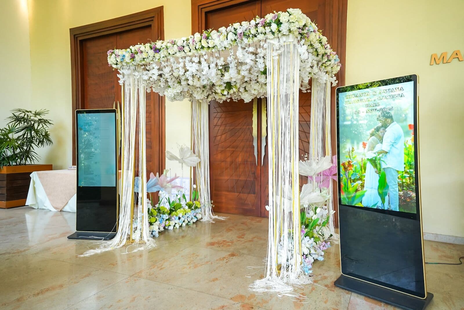 A flower canopy with hanging strings at a hall entrance, beside two welcome screens