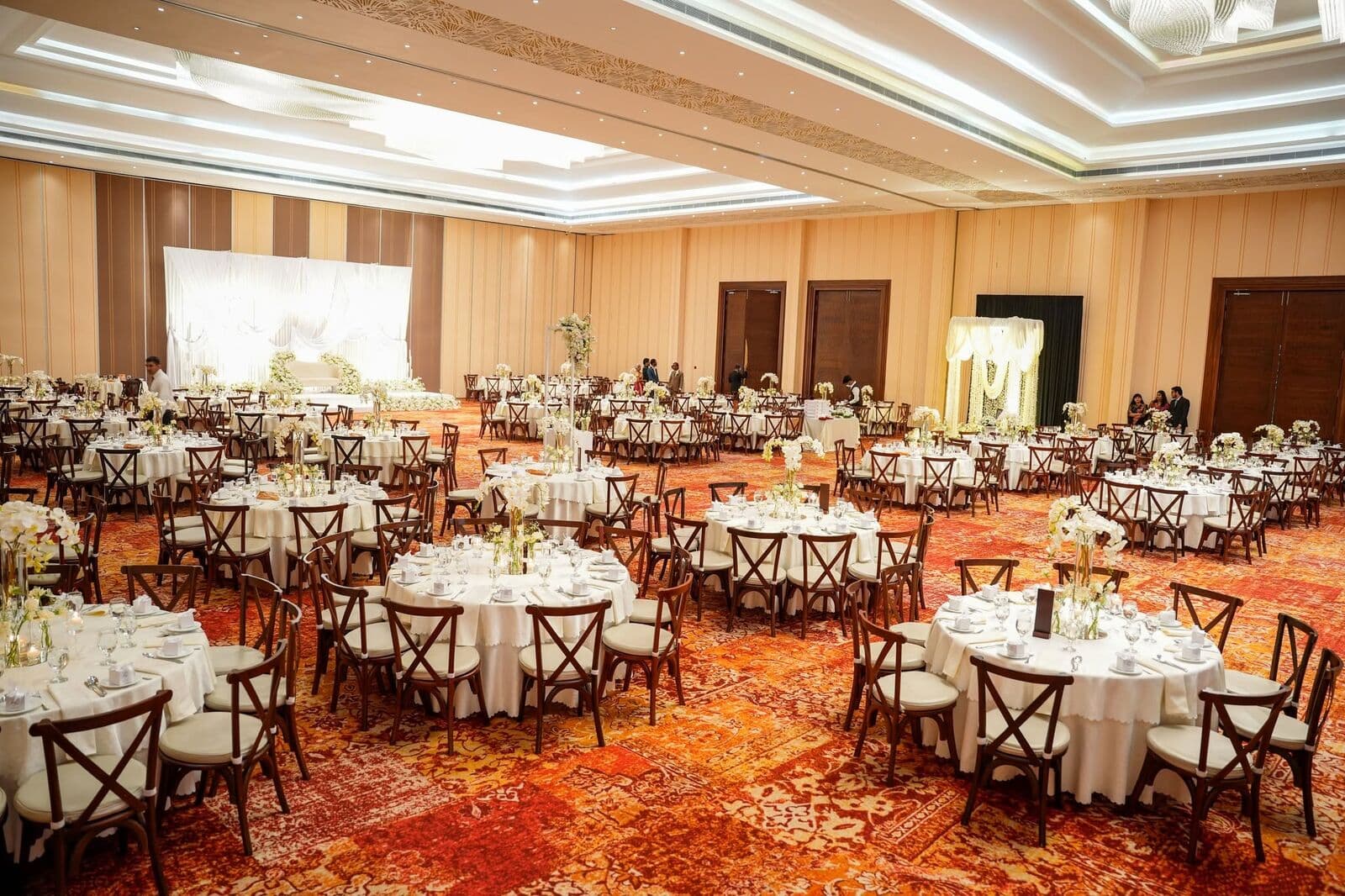 A hall laid for a wedding dinner: round tables in white linen, wooden chairs and a red patterned carpet