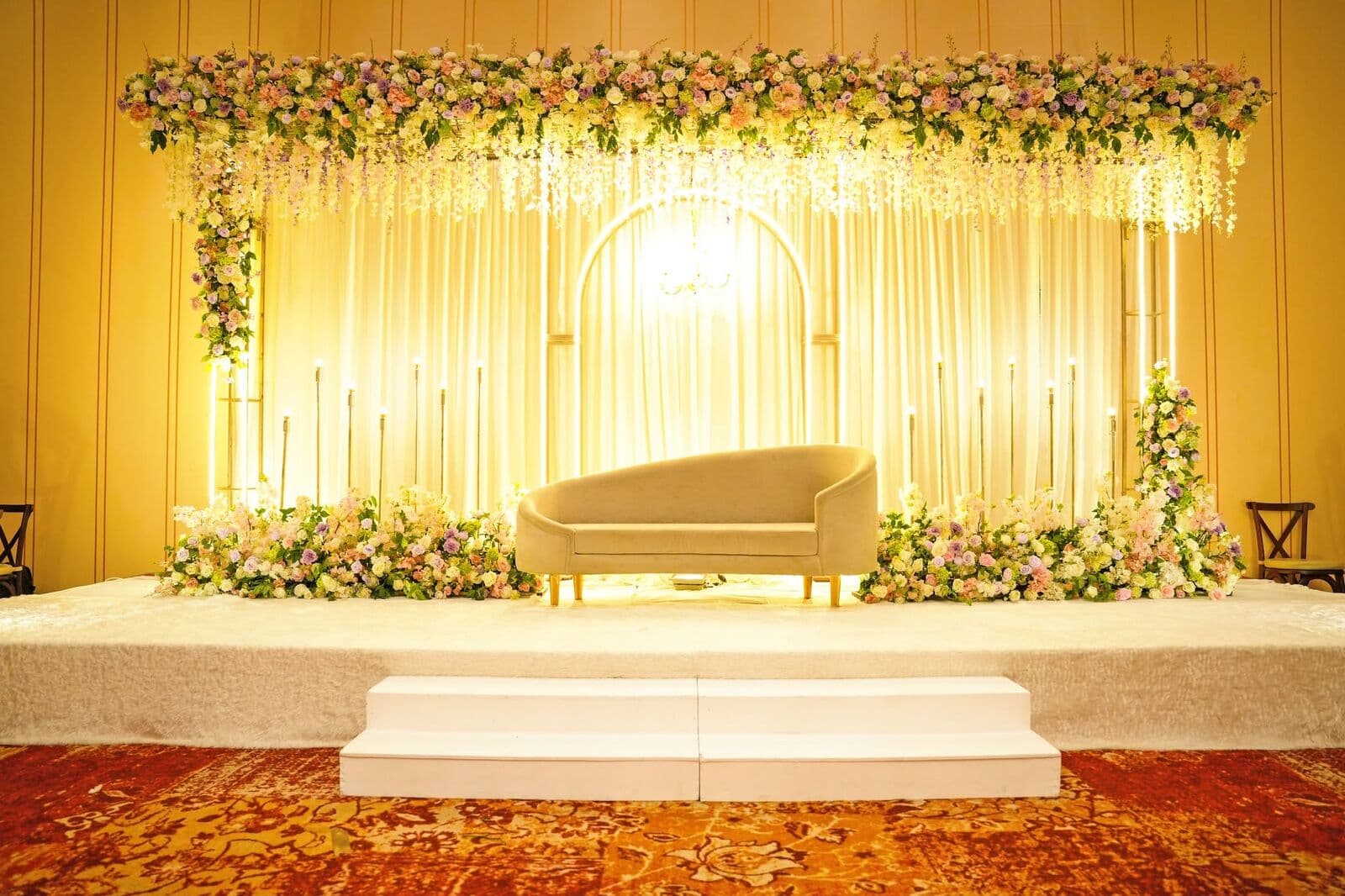 A stage lit in gold, with a sofa under an arch of flowers and candles