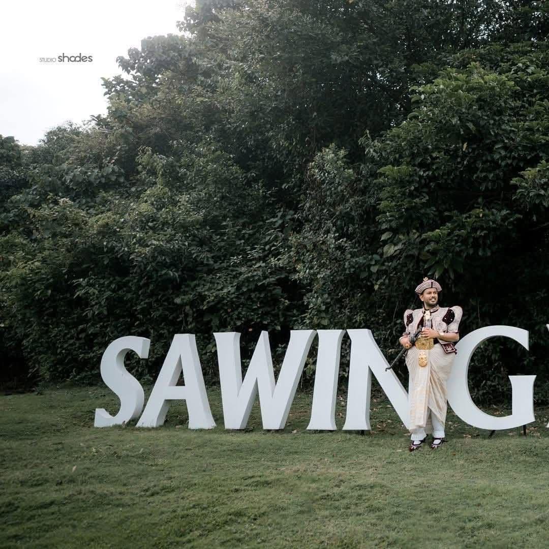 A groom in Kandyan dress leaning on the big white SAWINGIR letters on the lawn
