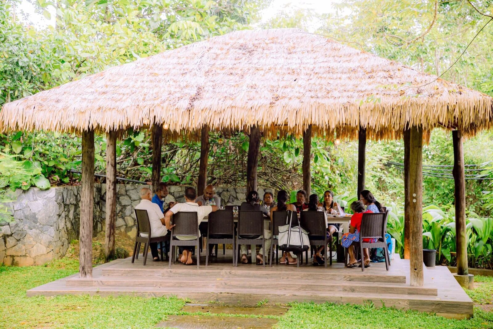 A family having lunch at a long table under a thatched hut in the garden