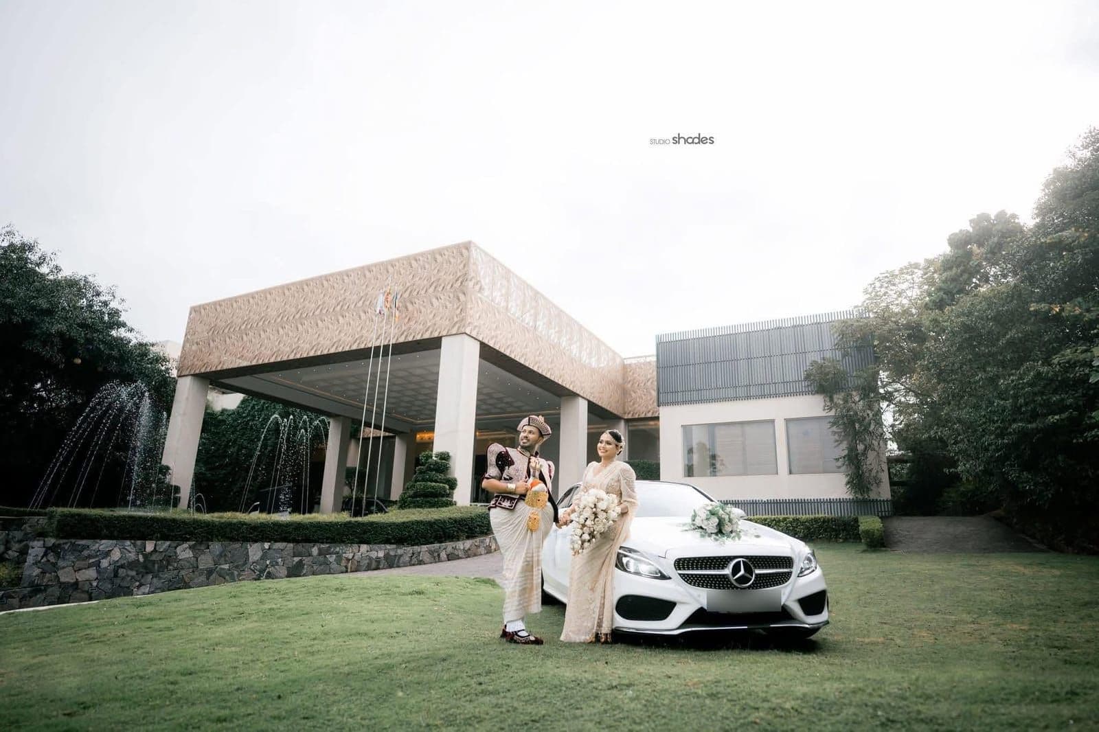 A couple in Kandyan wedding dress beside a flower-decked car on the lawn in front of the hotel entrance